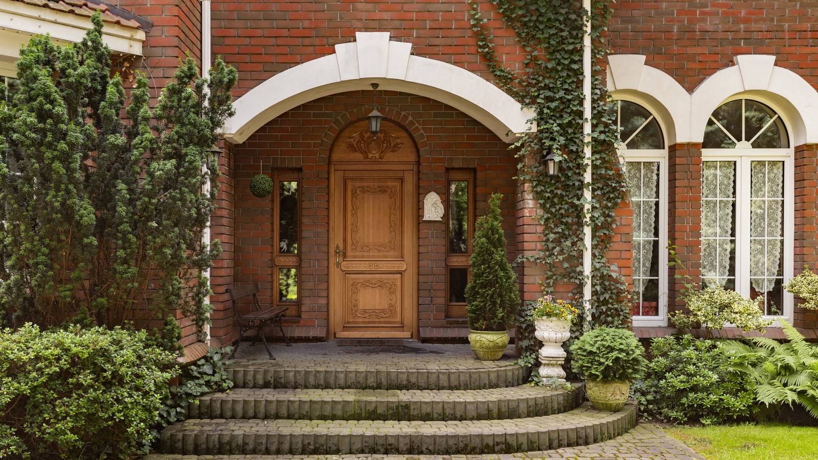 Plants, bushes and trees in front of wooden door of red brick mansion with windows